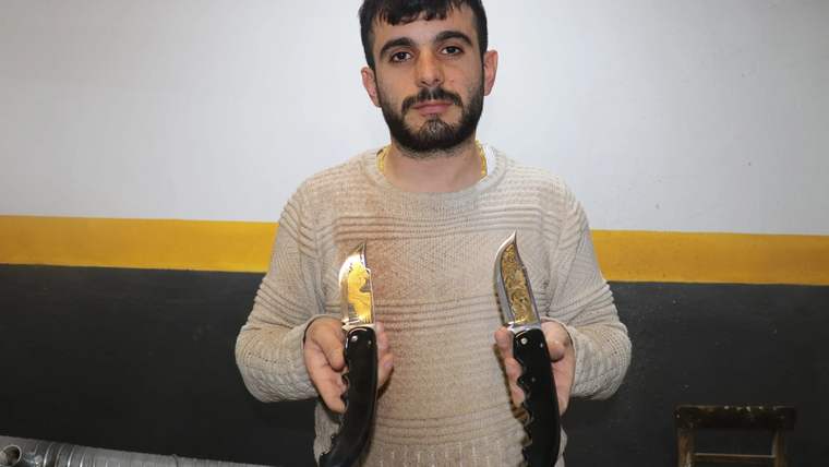 İki adet gravürlü ve altın kaplamalı bıçak tutan bir kişi, arka planda duvar görünmekte.