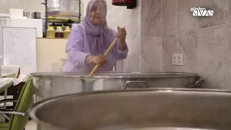 Büyük bir tencerede yemek karıştıran başörtülü bir kadın, mutfak ortamında çalışıyor.