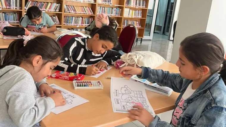 Kütüphanede bir masanın etrafında oturan çocuklar, kağıtlara boya kalemleriyle çizimler yapıyor.