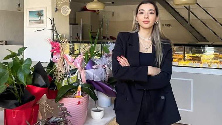 Kafede siyah kıyafetli bir kadın, çiçek aranjmanları ve vitrinlerin önünde kollarını bağlayarak duruyor.