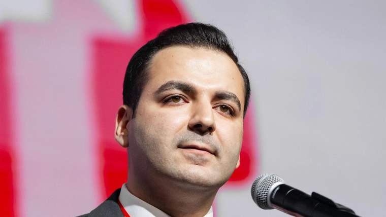 Kürsüde mikrofon önünde duran takım elbiseli bir kişi, boynunda kırmızı yaka kartıyla görülüyor.