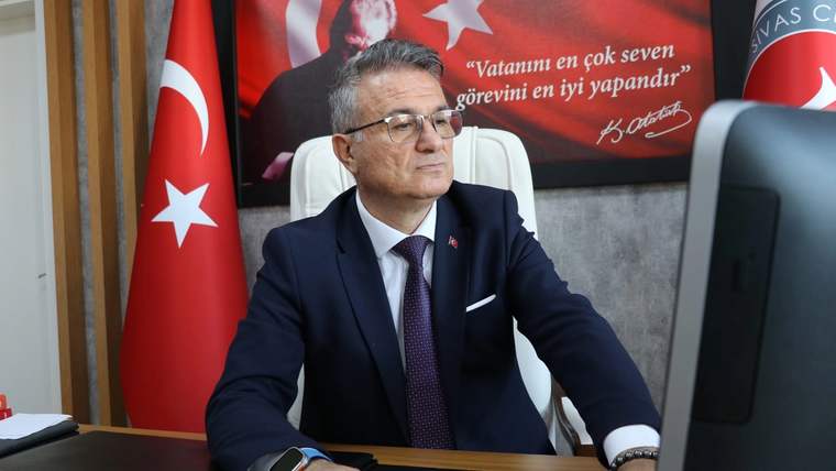 Bir kişi, masasında bilgisayar kullanarak çalışıyor; arka planda Türk bayrağı ve bir pankart var.