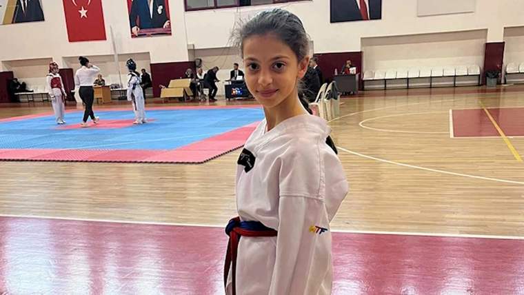 Beyaz tekvando giysisi giymiş, mavi kuşak takan bir kız çocuğu, spor salonunda gülümsüyor.