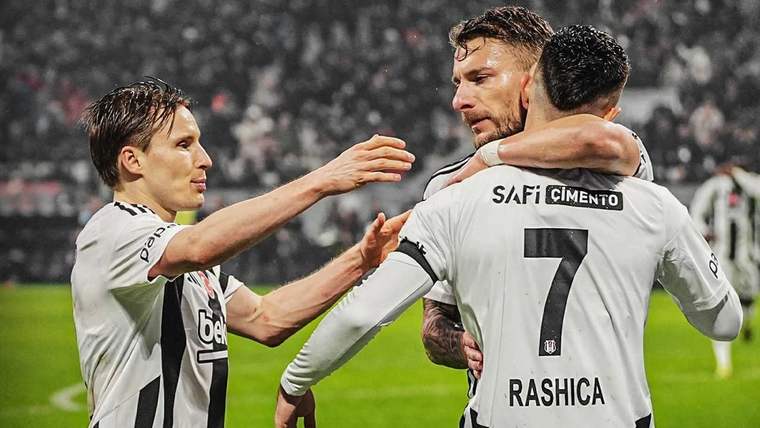 Üç futbolcu, sahada beyaz formalarıyla birbirine sarılırken arkadaki tribünler görülüyor.