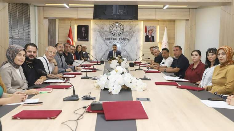 Toplantı salonunda uzun masa çevresinde oturan kişiler ve masada açık dosyalar görülüyor; duvarda kurum yazısı bulunuyor.