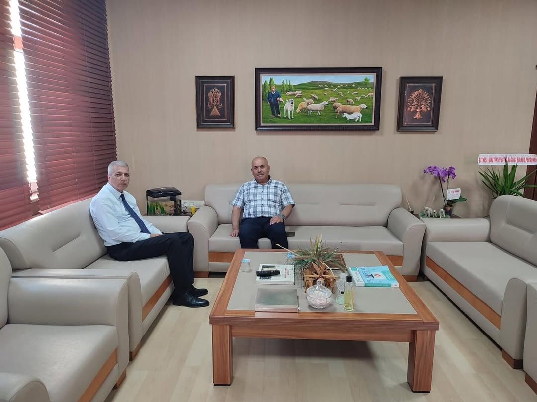 Ofis ortamında iki kişi koltuklarda oturuyor; önlerinde sehpa, üzerinde evraklar ve dekoratif eşyalar bulunuyor.