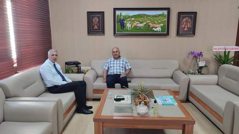 Ofis ortamında iki kişi koltuklarda oturuyor; önlerinde sehpa, üzerinde evraklar ve dekoratif eşyalar bulunuyor.