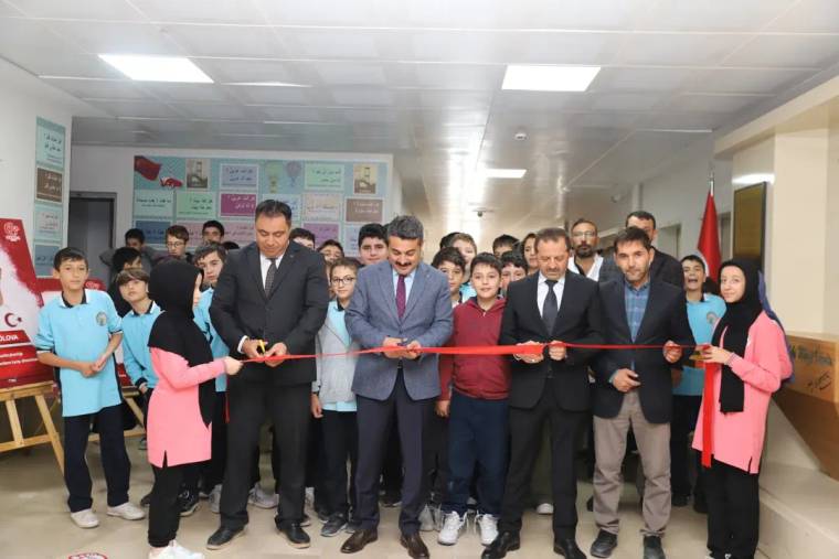 Okul koridorunda öğrenciler ve yetişkinler, kırmızı kurdeleyi makaslarla keserken yan yana duruyor.