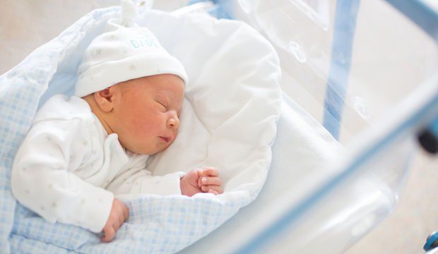 Beyaz bir örtü içinde uyuyan yeni doğmuş bir bebek, mavi bir bebek şapkası takıyor.
