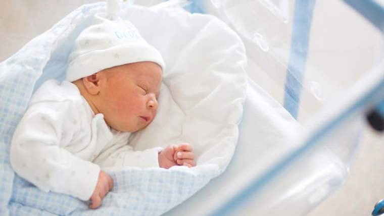 Beyaz bir örtü içinde uyuyan yeni doğmuş bir bebek, mavi bir bebek şapkası takıyor.