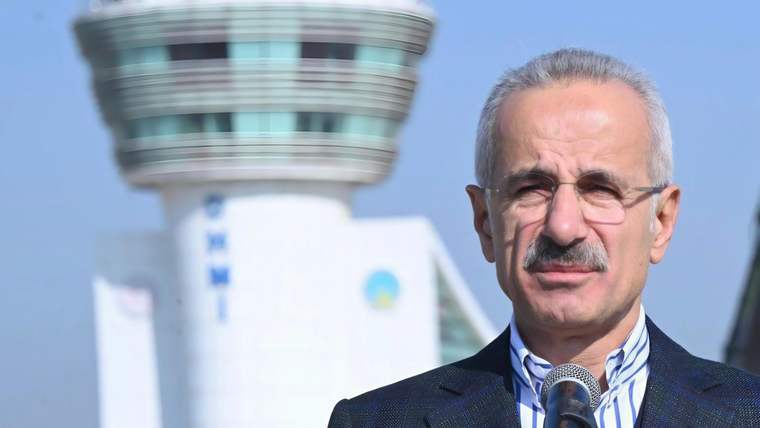 Mikrofon önünde konuşan gözlüklü bir erkek, arka planda havaalanı kontrol kulesiyle görülüyor.