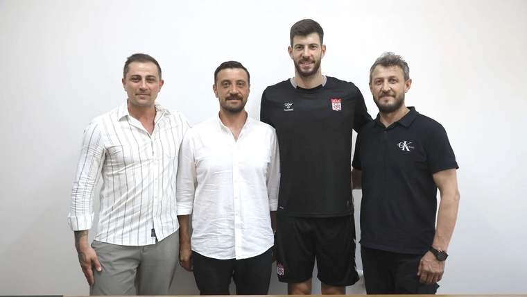 Beyaz duvar önünde duran dört erkekten biri siyah spor kıyafetleri, diğerleri günlük kıyafetler giyiyor.
