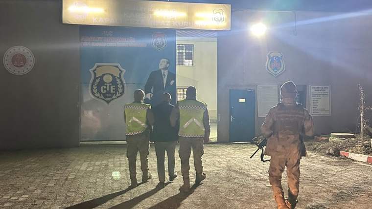 Gece aydınlatılan bir bina önünde, üç görevli bir kişiye eşlik ederken silahlı bir görevli bekliyor.