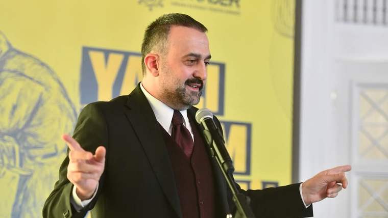 Takım elbiseli bir erkek, mikrofon önünde iki elini yana doğru uzatarak konuşurken görülüyor.
