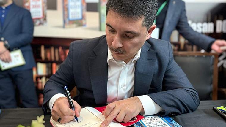 Bir adam, masanın üzerinde kitap imzalıyor. Arka planda kitap rafları görünmekte.
