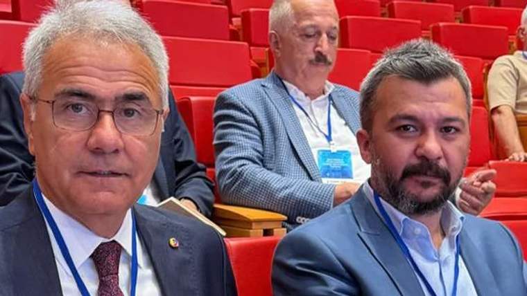 Bir toplantıda oturan iki kişi, arka planda başka katılımcılar var.