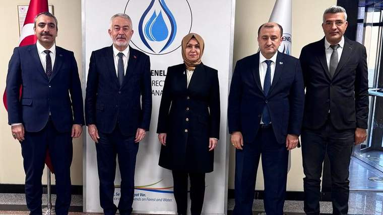Beş kişi, su damlası logosu ve yazıların bulunduğu pano önünde yan yana duruyor.