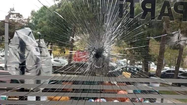 Çatlamış camın ardından, metal korkuluklar ve ağaçların yanında park edilmiş araçlar görünüyor.