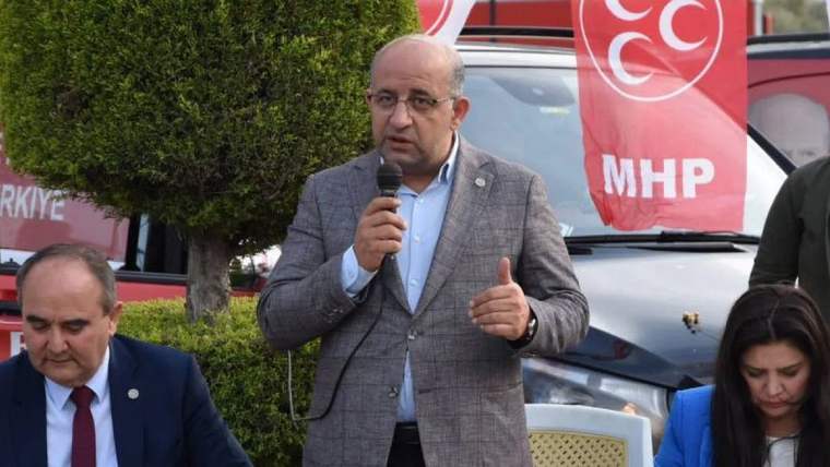 Mikrofonla konuşan bir kişi, arka planda MHP bayrakları ve bir araç görünmekte.