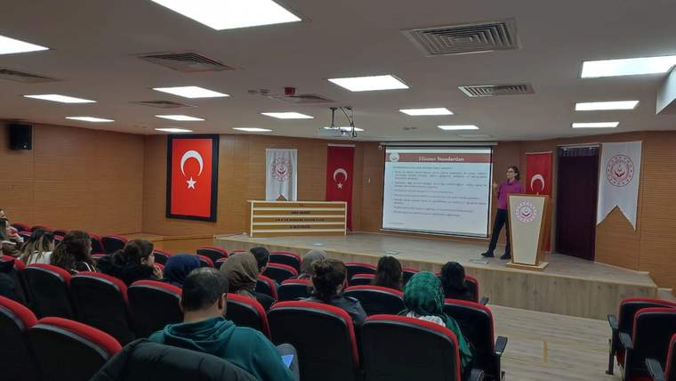 Konferans salonunda sıralara oturan dinleyiciler, kürsüde konuşan bir kişi ve sunum ekranı görülüyor.