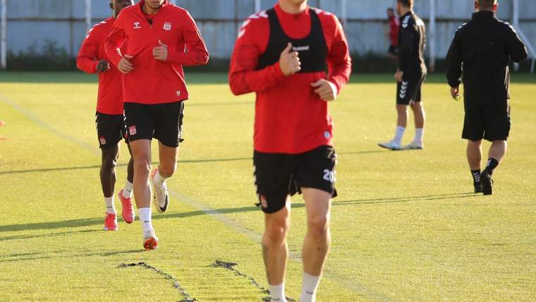 Çim sahada kırmızı antrenman formaları ve siyah şortlar giyen birkaç futbolcu koşu çalışması yapıyor.