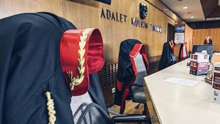 Mahkeme salonunda kürsü cübbeleri, masa ve duvarda “Adalet mülkün temelidir” yazısı görülüyor.