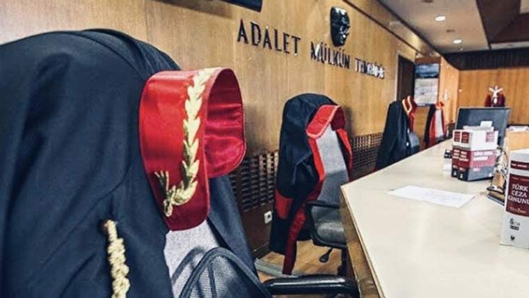 Duruşma salonunda cübbelerle örtülü sandalyeler ve üzerinde evraklar bulunan uzun masa görülüyor.