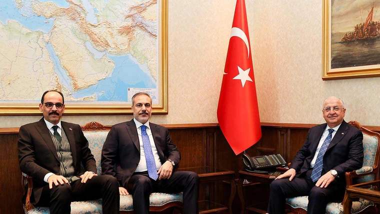 Duvarında harita ve Türk bayrağı bulunan bir odada üç erkek koltuklarda yan yana oturuyor.