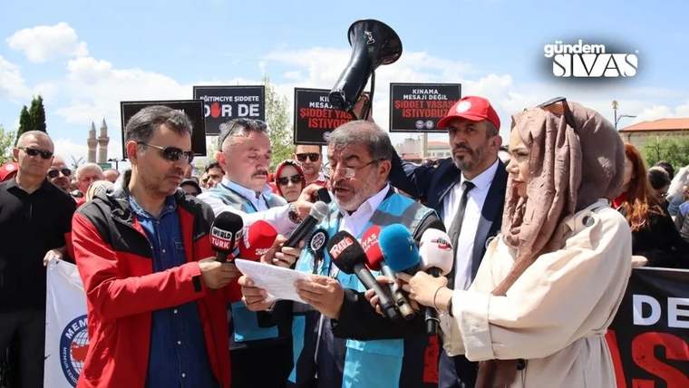 Mikrofonlarla çevrili bir kişi kâğıttan açıklama okurken çevresinde pankart taşıyan kalabalık görülüyor.