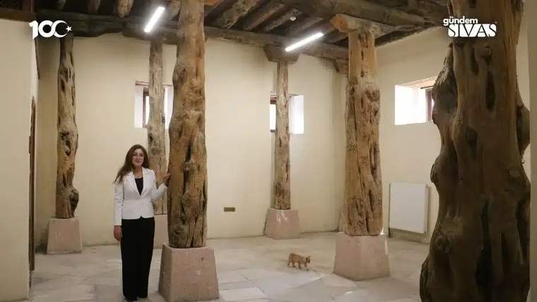 Tarihi bir konakta, ahşap sütunların arasında duran bir kadın ve yanındaki küçük bir kedi.