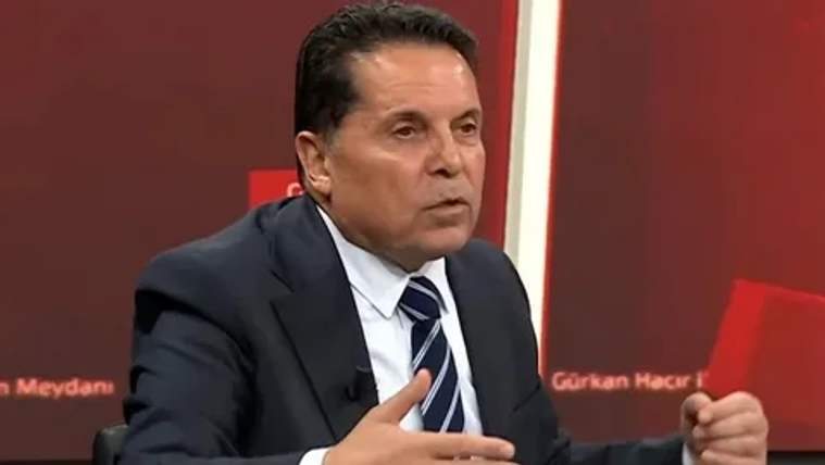 Takım elbiseli bir kişi, kırmızı arka plan önünde oturarak elleriyle konuşuyor.