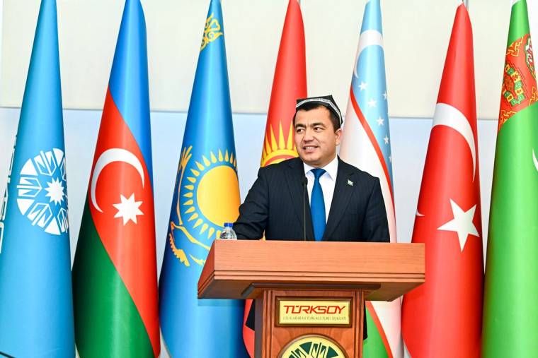 Bir kişi, üzerinde Türk bayrağı bulunan bir kürsüde konuşuyor; arkasında çeşitli ülke bayrakları yer alıyor.