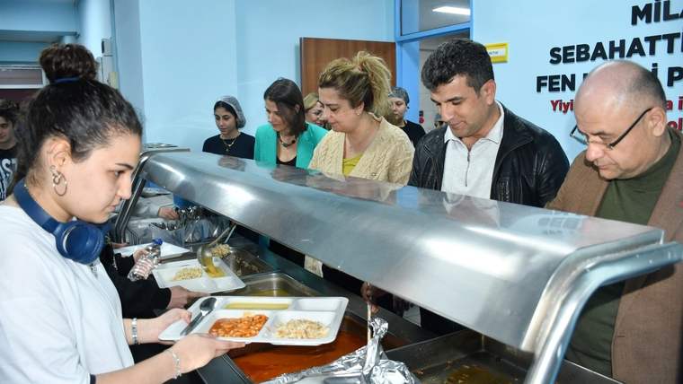 Yemekhanede bir öğrenci tepsiye yemek alırken, arkasında sırada bekleyen öğrenciler ve yetişkinler görülüyor.