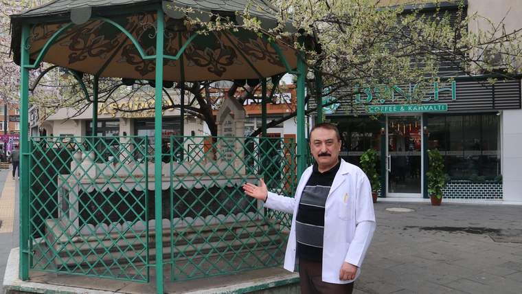 Kaldırımın ortasında yer alan bir türbe, etrafında yeşil demir parmaklıklar var. Türbenin önünde beyaz önlük giymiş bir kişi duruyor.