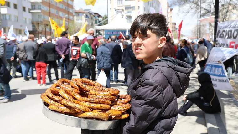 Bir çocuk, tepside simit taşırken kalabalık bir etkinlik arka planda yer alıyor.