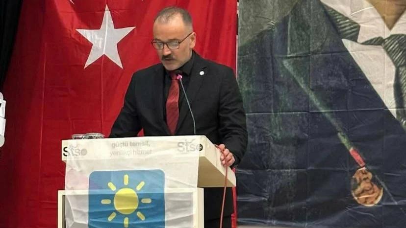 Kürsüde konuşan bir kişi, arkasında Türk bayrağı ve parti logosu bulunan bir pankart var.