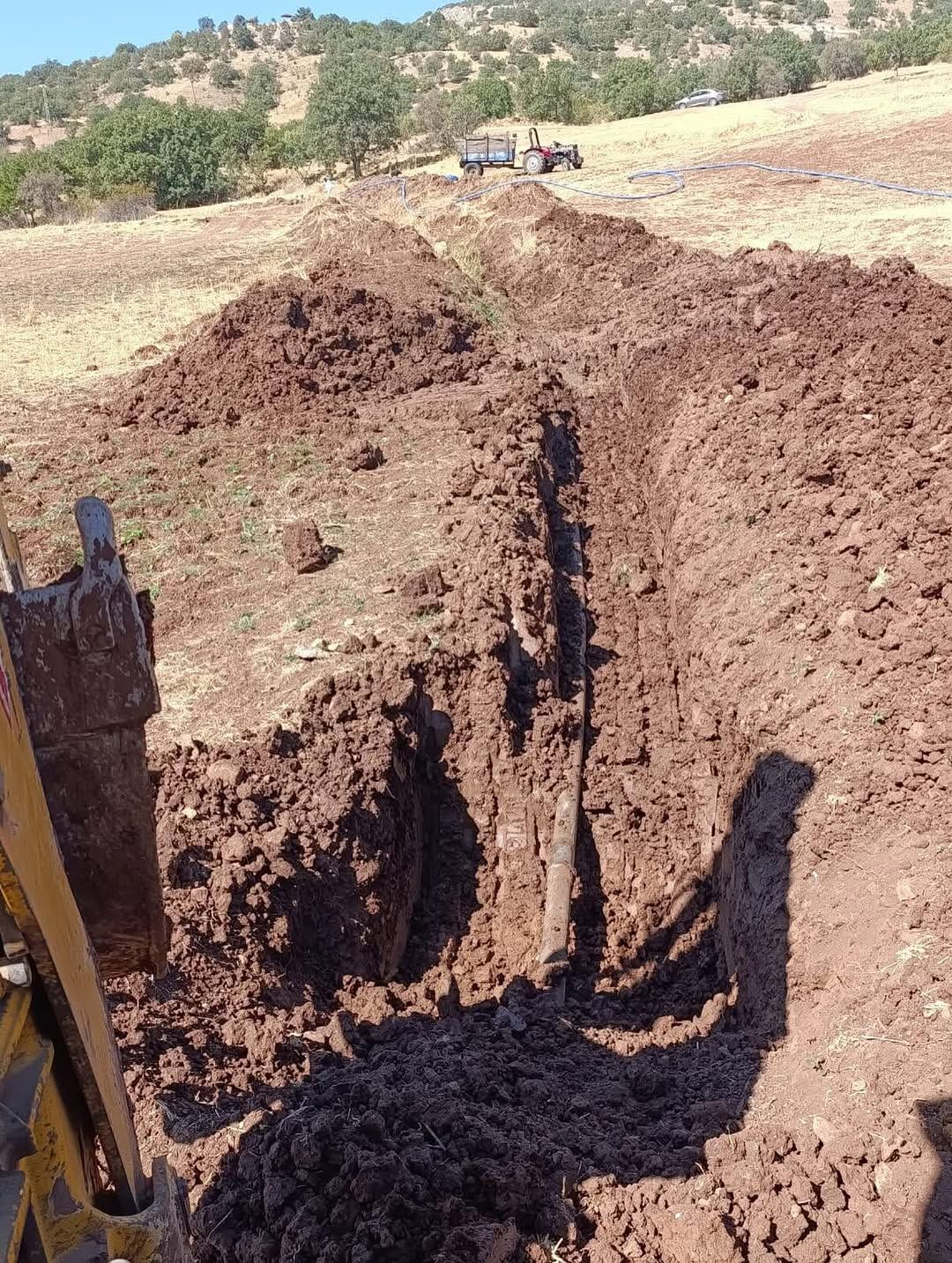 Toprak zeminde açılmış dar bir kanalda boru uzanıyor; sol kenarda iş makinesinin kepçesi görülüyor.