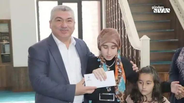 Bir adam, başörtülü genç kıza zarf uzatıyor; yanlarında küçük bir kız ve başka kişiler duruyor.