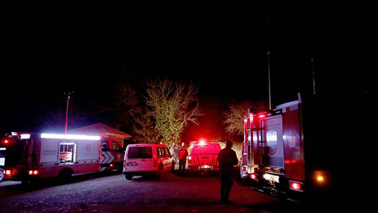 Gece karanlığında, ağaçlık alanda ambulans, kırmızı acil durum araçları ve birkaç kişi görülüyor.