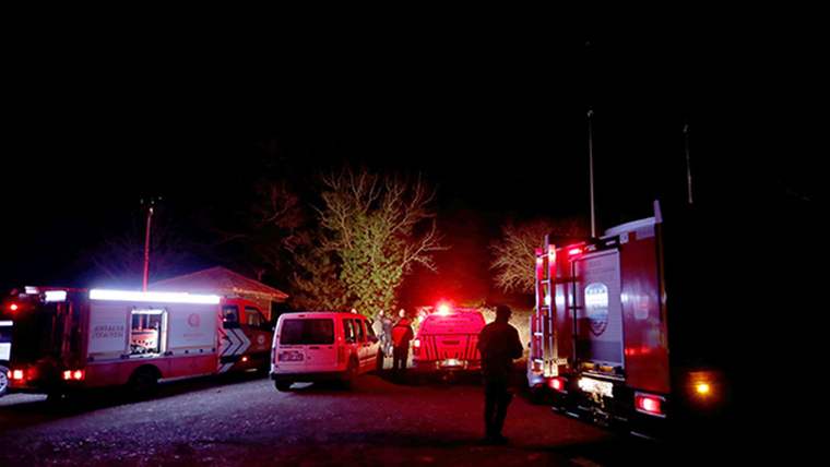 Gece karanlığında, ağaçlık alanda ambulans, kırmızı acil durum araçları ve birkaç kişi görülüyor.