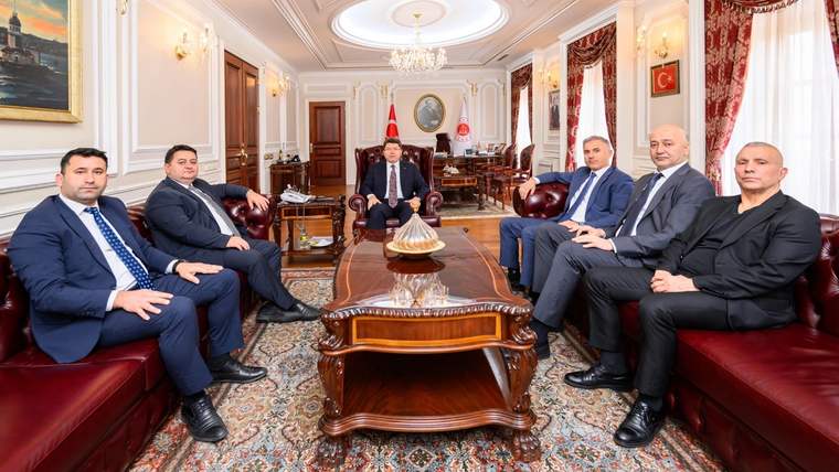 Resmî dekorasyonlu bir odada, ortada ve koltuklarda oturan altı kişi ile Türk bayrağı görülüyor.
