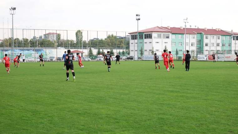 Yeşil futbol sahasında kırmızı ve siyah formalı oyuncular karşılaşma sırasında mücadele ediyor.