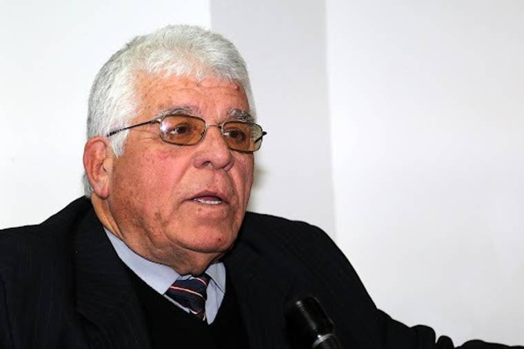 Gözlüklü, beyaz saçlı bir kişi takım elbiseyle otururken önünde mikrofon görülüyor.