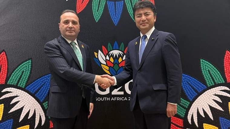 Takım elbiseli iki erkek, üzerinde renkli desenler ve “SOUTH AFRICA” yazısı bulunan panonun önünde tokalaşıyor.