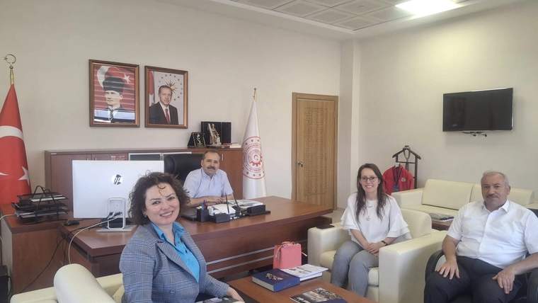 Bir ofiste, masanın etrafında oturan dört kişi, arka planda Türk bayrağı ve Atatürk portresi var.