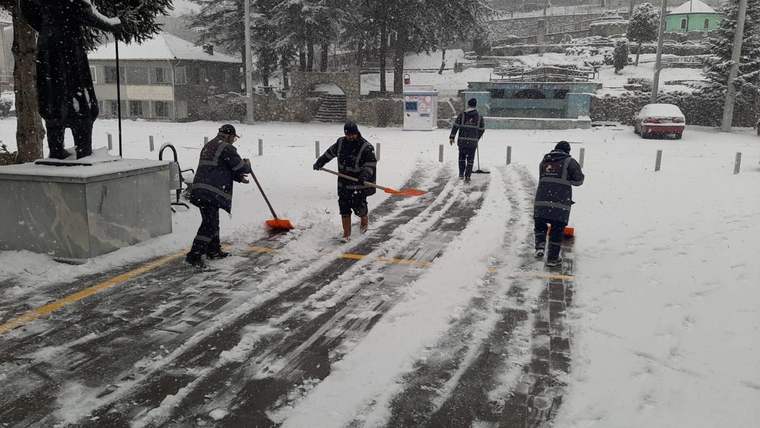 Kar yağışı altında dört görevli, kürek ve fırçalarla yokuş yoldaki karı temizliyor.