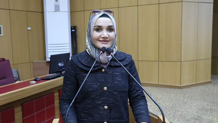 Başında gözlük bulunan başörtülü bir kadın, ahşap panelli salonda iki mikrofon önünde konuşuyor.