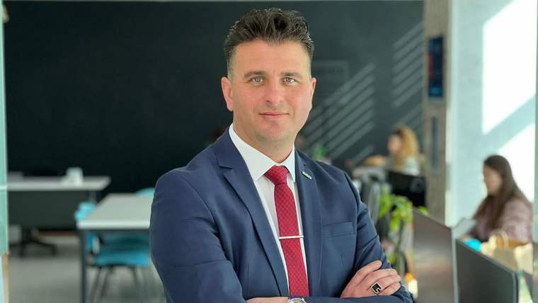 Lacivert takım elbiseli, kırmızı kravatlı bir kişi kapalı bir mekânda kollarını bağlayarak duruyor.