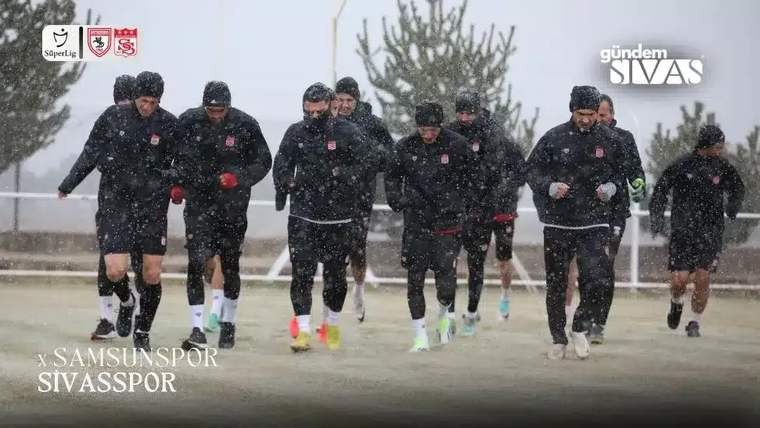 Kar yağışı altında siyah antrenman kıyafetleri giyen futbolcular, açık alanda birlikte koşuyor.