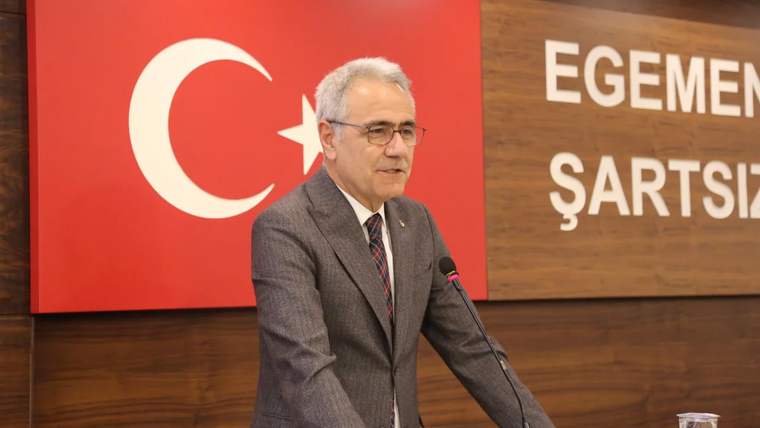Kırmızı-beyaz Türk bayrağının önünde, bir masanın arkasında konuşan bir kişi.
